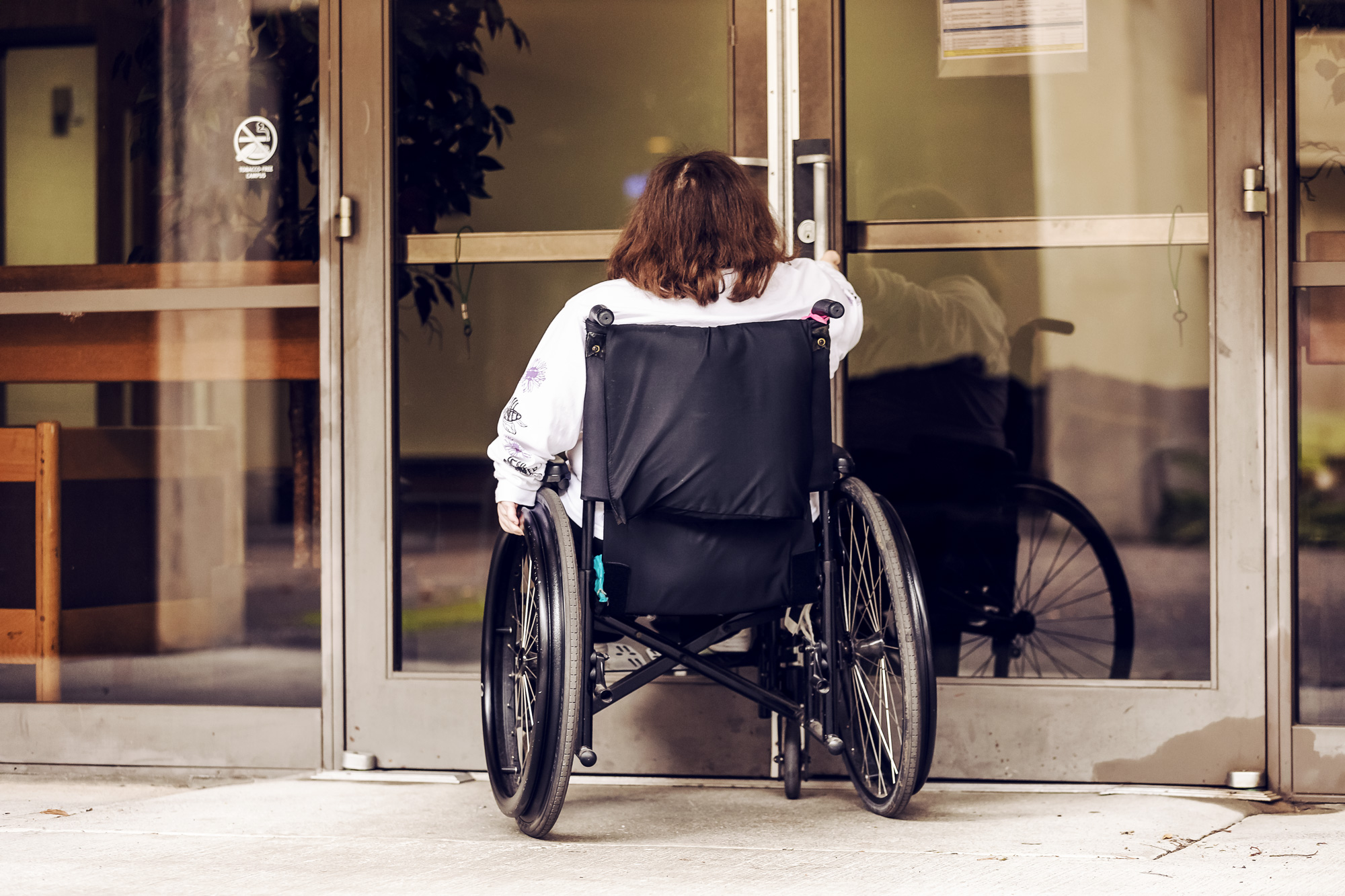 Photo capturing someone in a wheelchair trying to pull open non-automatic doors to show accessibility issues on the campus.