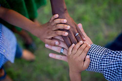 Web photograph of multiple hands reaching together with grass in the background.