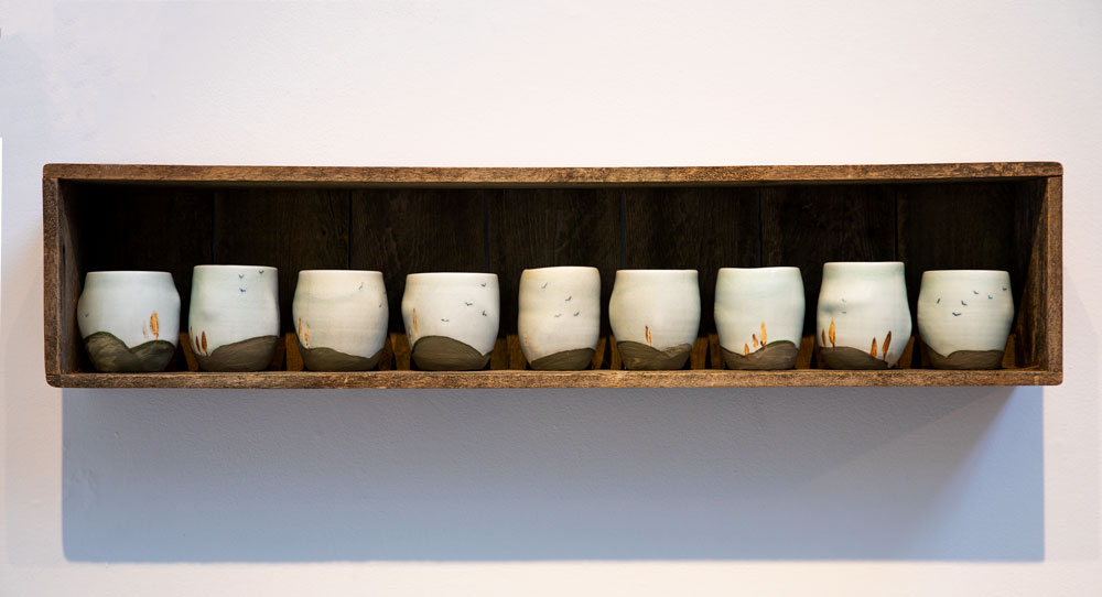 Photograph of 9 ceramic cups lined up on a wooden shelf, each with a simple hilly scene on the bottom and some birds flying in the sky.