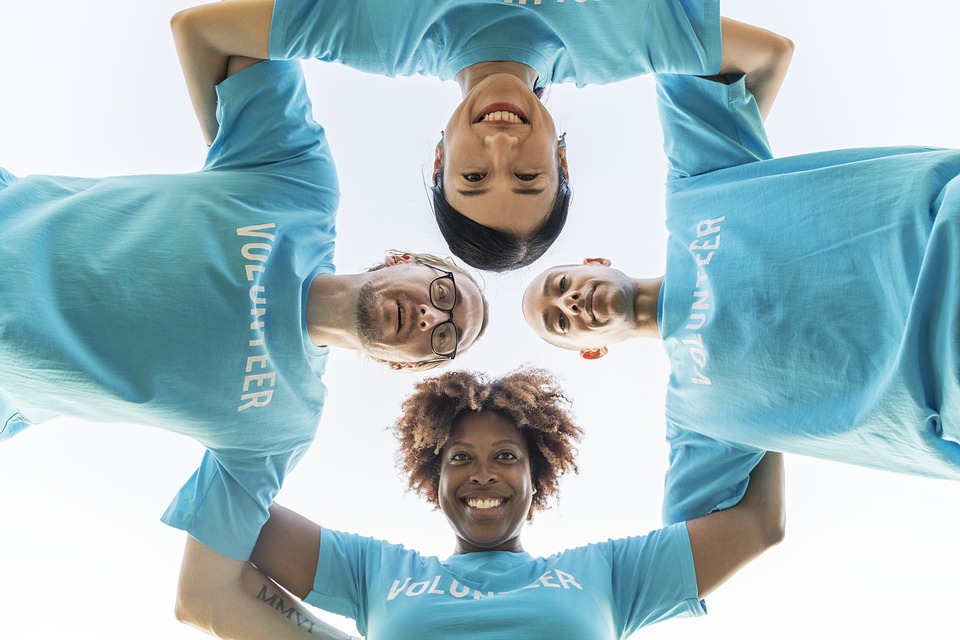 Web photograph of four individuals with 'VOLUNTEER' on their t-shirts, standing with heads together in a circle, camera angle point up at them.