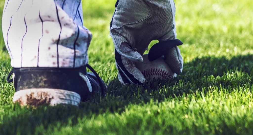 A close up of a baseball player's hand on the ground picking up a baseball in the grass.