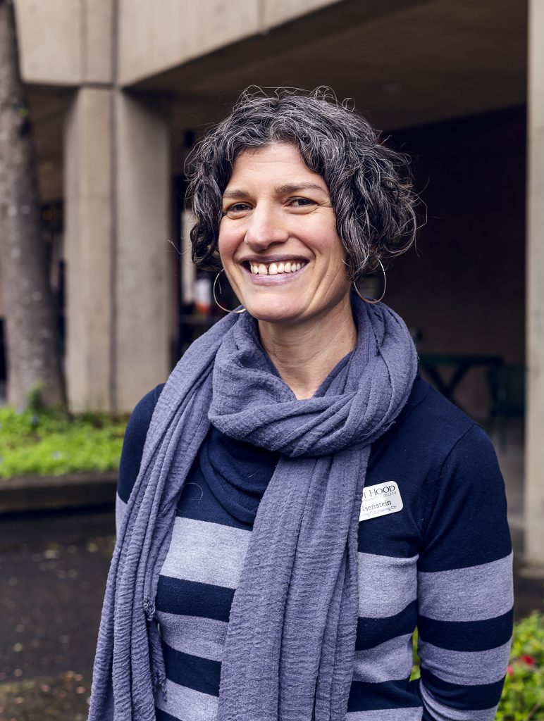 Adult woman with short dark curly hair, light skin, and a big smile posing for photo with arms behind back. Wearing long sleeve striped shirt and a scarf outside.