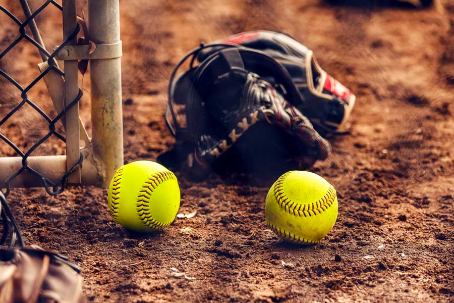 A close up of a softball mitt in the dirt with two softballs beside it.