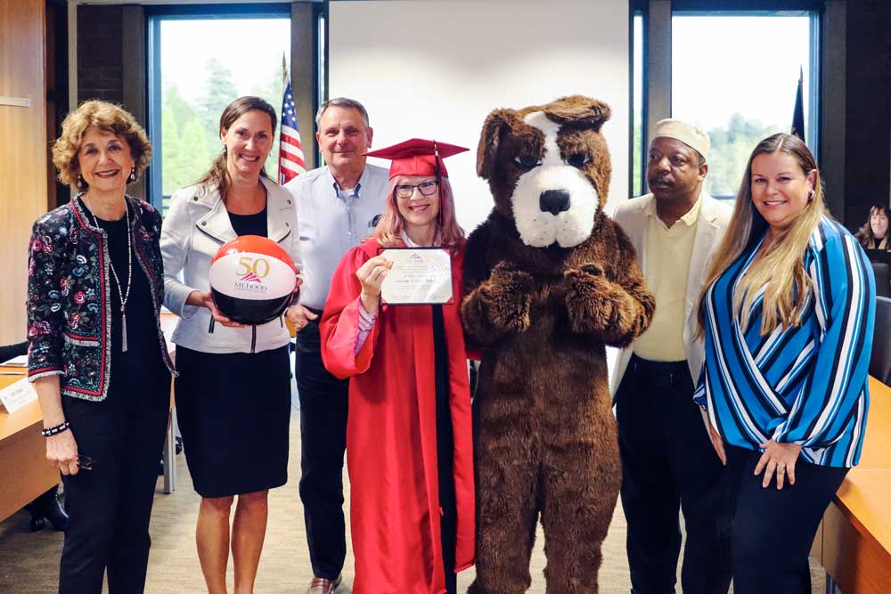 The board of education posing with Annette Mattson in the center wearing a cap and gown while holding her degree. Mt. Hood's mascot stands to the left of Mattson.