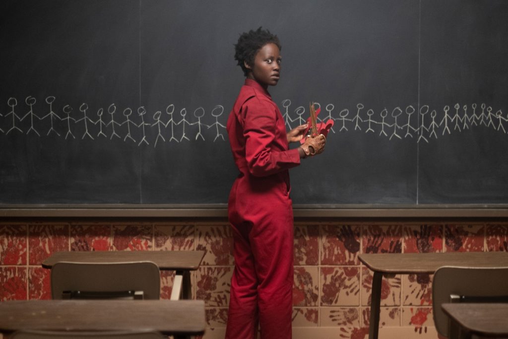 A woman in a red jumpsuit standing at the front of a classroom with stick figures drawn on a chalkboard.