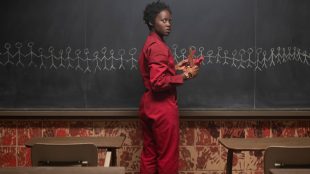 A woman in a red jumpsuit standing at the front of a classroom with stick figures drawn on a chalkboard.