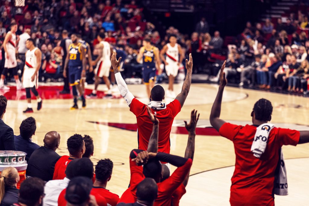 Photo at the Moda Center court with Blazers sideline players reacting to a play, back turned to camera.