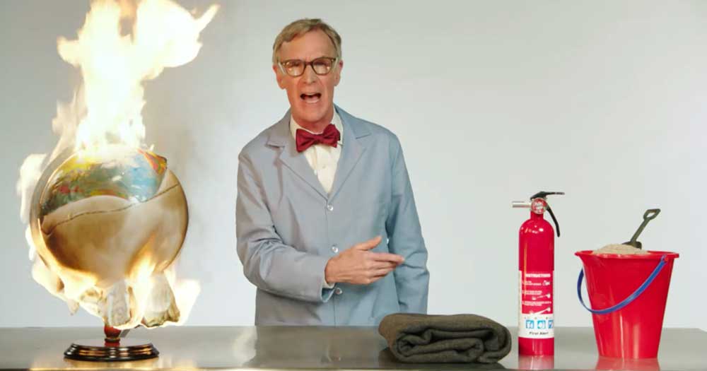 Bill Nye yelling as a globe is on fire on a table with a blanket, fire extinguisher, and blanket, not being used.
