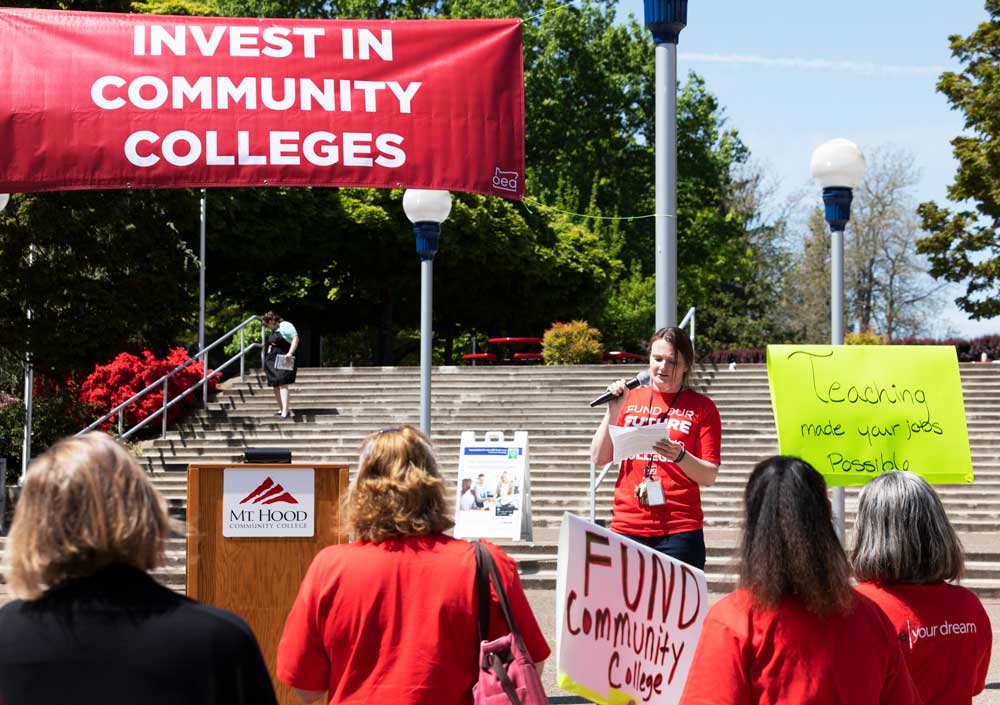 A banner states "Invest in Community Colleges" in the background with Cheryl facing the crowd speaking in a microphone. Two signs are visible. One says "fund community college" the other says "Teaching made your jobs possible."