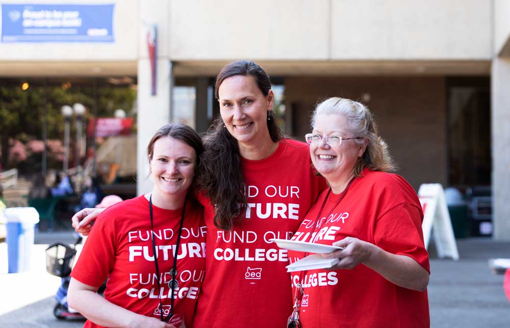 Three women pose for a photo, all wearing the same red shirt which states, "Fund our future, fund our colleges."