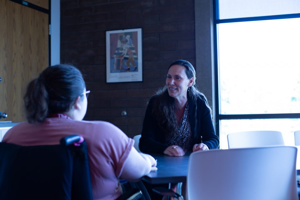 Lisa Skari sitting at the table in her office smiling while she talks to reporter Cassie Wilson. Photo taken from behind Cassie looking at Lisa.