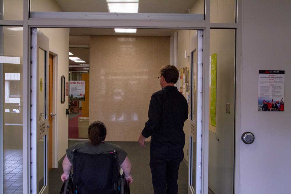 Young woman wheels herself in wheelchair through MHCC library doors next to Dave Pontious from Accessible Education Services