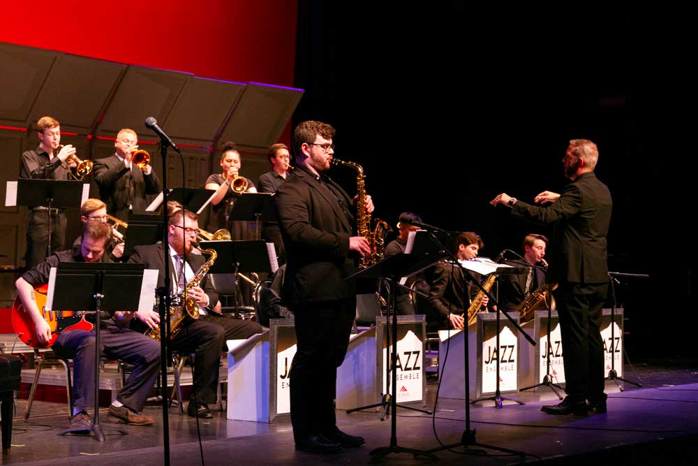 All the jazz band members playing their instruments with one saxophone player standing at the front for a solo while the director conducts everyone through the music.